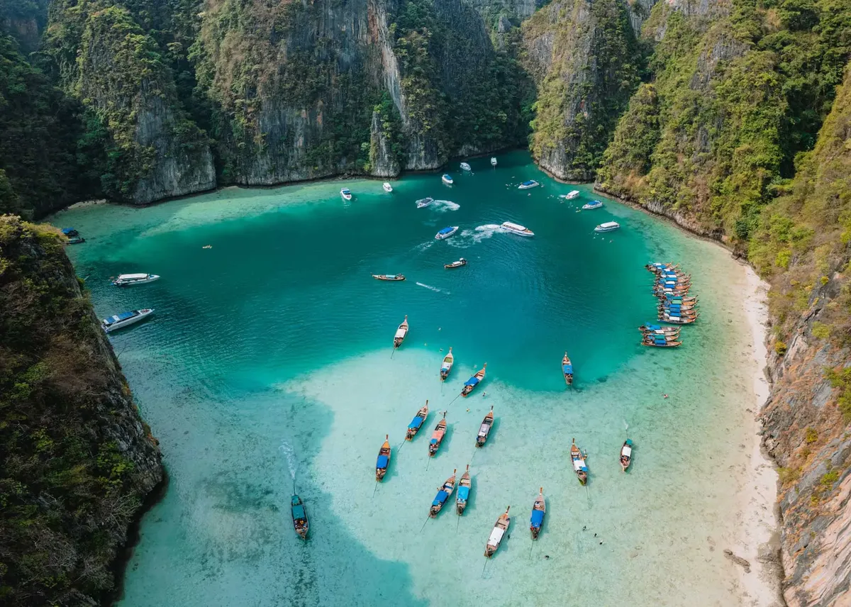 Aerial view of a Maya bay, Phuket with clear turquoise water, surrounded by steep green cliffs and numerous boats anchored near the sandy shore.