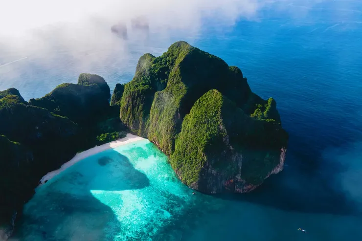 Aerial view of a secluded Maya Bay with steep, green cliffs, a white sandy beach, and clear turquoise water surrounded by the ocean.